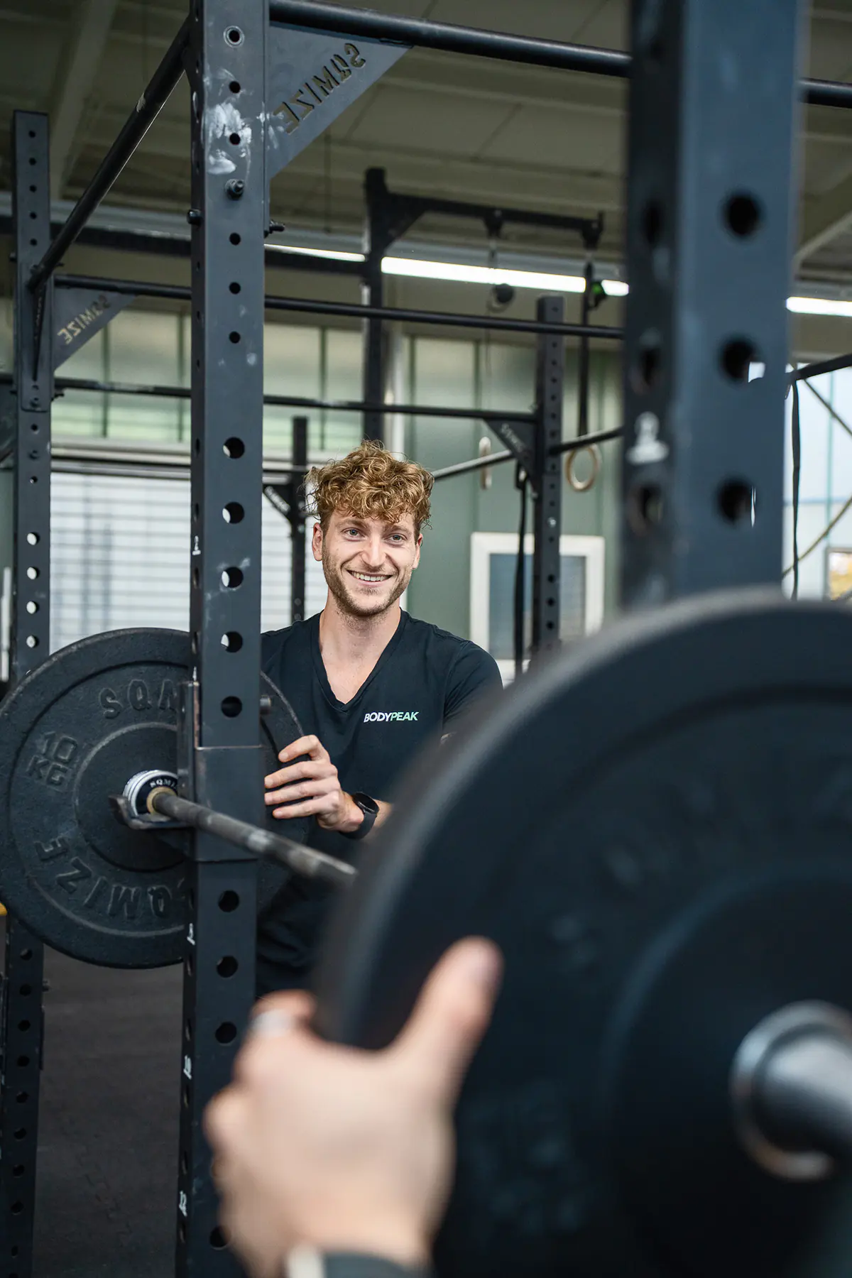 Patrick Keicher im schwarzen BodyPeak-Shirt lehnt lächelnd an einer Langhantel im Rack